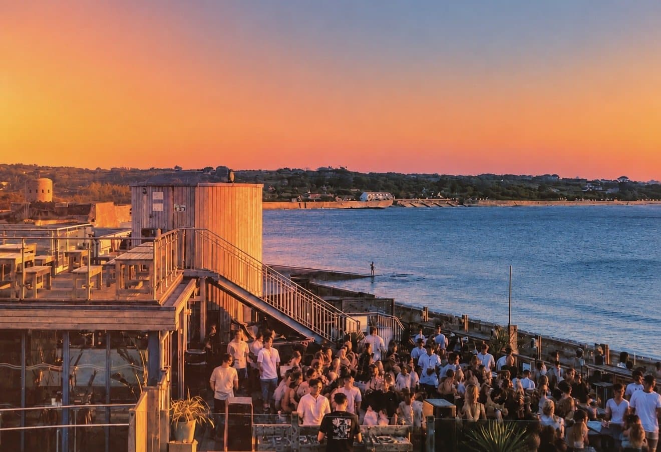 Sunset over Vazon Bay from the Vistas roof terrace, with a busy crowd on the deck and the sea beyond